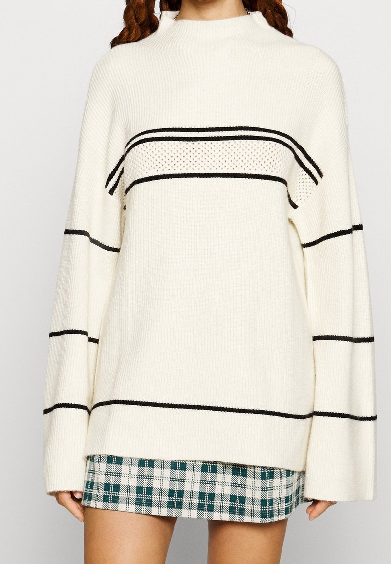 Jersey crema de gran tamaño con rayas horizontales negras, textura acanalada y cuello alto. Combinado con una falda mini de cuadros verde y blanca.