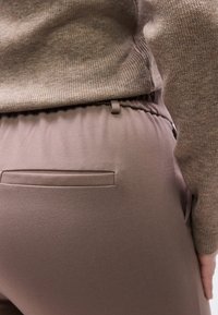 Κοντινό πλάνο ενός ατόμου που φοράει παντελόνι ταφέ με ελαστική μέση και πίσω τσέπη, καθώς και καφέ πλεκτό πουλόβερ.