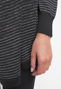 Main reposant à côté du corps portant un pull à manches longues rayé gris foncé et noir et un pantalon noir.