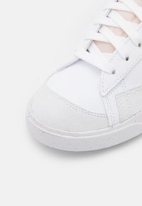 Hvit sneakers med en teksturert semsket tærkappe, glatt skinnøvre del, flate hvite snøringer og en hvit flekket såle.