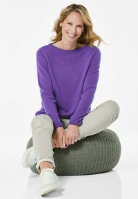 Lila Strickpullover, runder Halsausschnitt, lockere Passform. Modell sitzt auf einem strukturierten runden grauen Pouf, trägt hellbeige Hosen und weiße Sneakers.