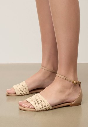 Jalad, mis kannavad beežikaid lamedaid sandaale, millel on pahkluu rihmad ja keerukad kootud esiosad, seisavad siledal pinnal.