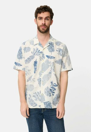 Mężczyzna w kremowej koszuli na krótki rękaw z niebieskimi abstrakcyjnymi wzorami liści i roślin, ubrany w ciemnoniebieskie dżinsy, stojący na białym tle.