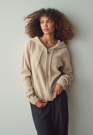 Beige ziphoodie van zacht materiaal met een capuchon, geribde manchetten en zoom, gedragen over donkere broeken. Model poseert tegen een lichte achtergrond.