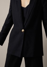 Dunkelblaues Sakko aus strukturiertem Stoff, mit einem einzelnen runden goldenen Knopf und tailliertem Schnitt, darunter ein Top mit tiefem V-Ausschnitt.