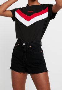 Camiseta de algodón negra con patrón de chevron rojo y blanco; combinada con pantalones cortos de mezclilla negros de tiro alto con un dobladillo sin terminar y botón frontal.