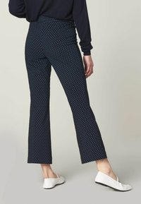 Kvinna i marinblå cropped-byxor med små vita prickar och vita platta skor, stående mot en enkel ljus bakgrund.