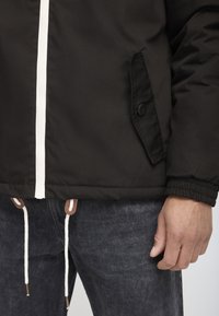 Black jacket with a white zipper, side pocket with button, elastic cuffs, and drawstrings at the hem. Worn over dark denim jeans.