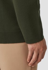 Groene gebreide trui met geribde manchetten, getoond naast een beige stof, die de textuur en kleurcontrast benadrukt.