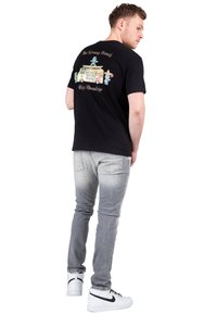 Sort bomulds t-shirt med et farverigt grafisk tryk af en varevogn og tegneseriefigurer på ryggen, parret med lysegrå jeans og hvide sneakers.