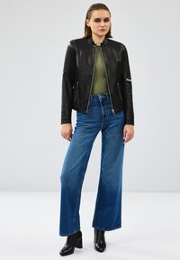 Chaqueta de cuero negra con hombros texturizados, cierre con cremallera en el frente, combinada con vaqueros de tiro alto de pierna ancha azules y botas negras de tobillo.