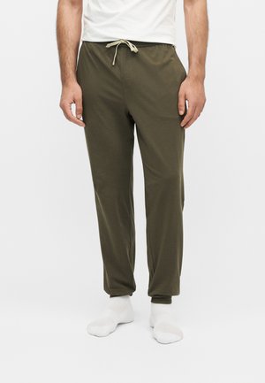 Man draagt olijfgroene broek met trekkoord en witte sokken, staand tegen een effen witte achtergrond.