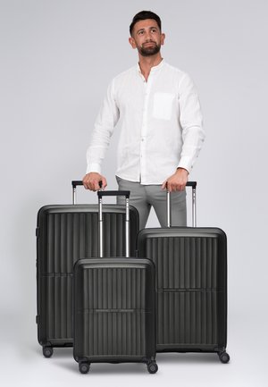 Trois valises rigides noires avec une texture à nervures verticales, des poignées en aluminium et des roues. Différentes tailles présentées devant un fond clair.
