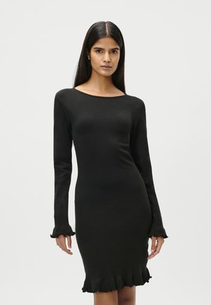 Femeie cu păr lung și închis la culoare, purtând o rochie neagră mulată cu mâneci lungi și volane la manșete și tiv, stând în fața unui fundal gri deschis.