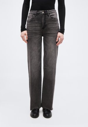 Persoană care poartă blugi negri decolorați, cu talie înaltă și croială dreaptă, cu pantofi negri și un top negru cu mâneci lungi, mulat, pe un fundal simplu.