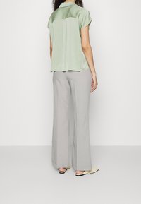 Ljusgrön satinblus med korta ärmar, detalj med veck bak, tillsammans med ljusgrå byxor med vida ben och beige slipskor.