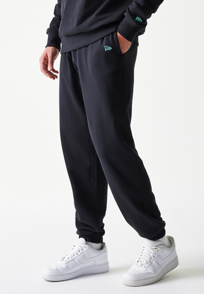 Μαύροι φόρμες με ελαστικά μανσέτες και τσέπες, κατασκευασμένοι από μαλακό ύφασμα· διαθέτουν μικρό τουρκουάζ λογότυπο στο αριστερό μηρό. Συνδυασμένοι με λευκά αθλητικά παπούτσια.