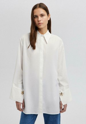 Młoda kobieta ubrana w oversize'ową białą koszulę z szerokimi mankietami i spinkami w kształcie gwiazdek, w połączeniu z niebieskimi jeansami, stojąca na jasnym tle.