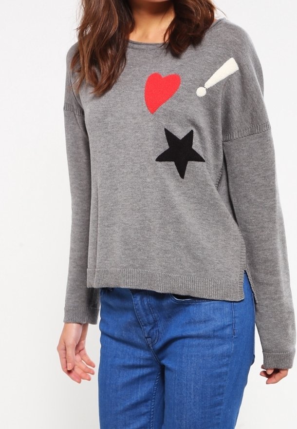 Femme portant un pull gris avec des motifs de cœur rouge, d'étoile noire et de point d'exclamation blanc, associé à un jean bleu.
