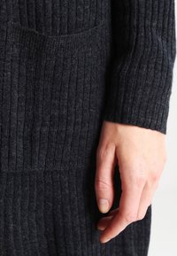 Ingrandimento di una mano poggiata accanto a un maglione a coste grigio scuro con una tasca visibile sul lato.