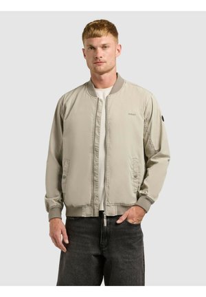 Mężczyzna w beżowej kurtce bomberce na białej koszuli i ciemnoszarych dżinsach, stojący z jedną ręką w kieszeni na jednolitym tle.