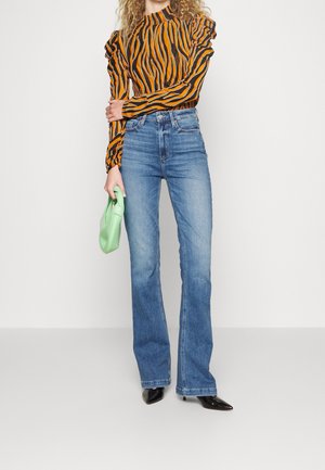 Oranje en zwart gestreepte lange mouw top met gerimpelde mouwen, gecombineerd met lichtblauwe flare jeans en een groene clutch. Zwarte puntige hakken.