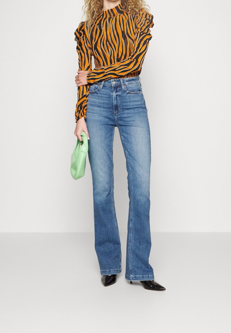 Top de manga larga a rayas naranjas y negras con mangas acampanadas, combinado con unos pantalones vaqueros de campana azul claro y un bolso de mano verde. Tacones negros de punta.