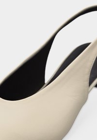 Μπεζ δερμάτινο γυναικείο παπούτσι με λουράκι πίσω, με μαύρη εσωτερική επένδυση, εμφανιζόμενες λεπτομέρειες πλευράς και λουριού πτέρνας σε λευκό φόντο.