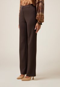 Braune maßgeschneiderte Hose mit geradem Bein, Knopfverschluss und glatter Textur. Kombiniert mit nudefarbenen High Heels.