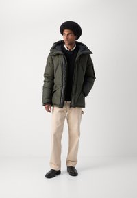 Chaqueta acolchada en verde oscuro con capucha, cierre frontal con cremallera y bolsillos laterales. Llevada sobre un suéter negro y pantalones beige, combinada con zapatos negros.