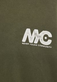 Lettres "NYC" blanches usées avec le texte "Never Young Community" en dessous sur un fond en tissu vert olive.