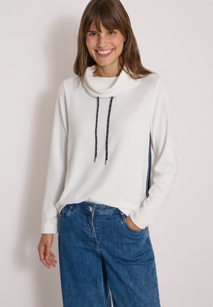 Jeune femme souriante portant un haut blanc à manches longues avec col cheminée, cordons noirs et jean denim bleu, debout devant un fond uni.