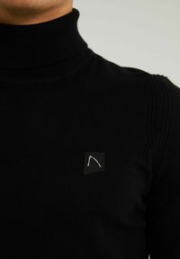 Schwarzer Rollkragenpullover mit gerippten Nähten an den Schultern und einem kleinen schwarzen Patch mit einem weißen geometrischen Design. Weicher, dehnbarer Stoff.