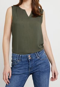 Olive green sleeveless blouse with a v-neck, made of smooth fabric. Paired with blue high-waisted jeans, featuring two front buttons.