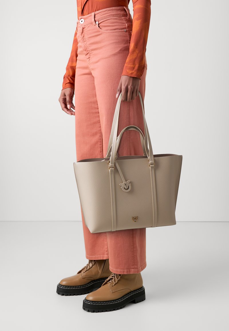 Bežová taška cez rameno s textúrovaným povrchom, dvojitými vrchnými uchami a dekoratívnym emblémom, ktorú drží osoba oblečená v ružových širokých nohaviciach a hnedých topánkach.