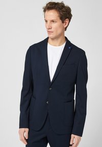 Marineblazer mit Reverskragen, zwei Vorderaschen, Ein-Knopf-Verschluss und kontrastierenden blauen Akzenten. Glattes, strukturiertes Material.