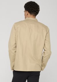 Veste en coton beige avec col, manches longues et coupe droite, présentant des détails de couture minimalistes et une texture lisse.