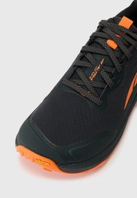 Chaussure de trail running noire avec tige en tissu texturé, accents orange et semelle extérieure Vibram. Dispose d'un système de laçage sécurisé pour un bon ajustement.