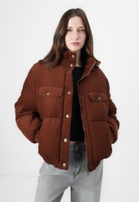 Bruine pufferjack met een corduroy textuur, voorzien van drukknoopjes, twee borstzakken en elastische manchetten. Draagbaar over een zwarte top.