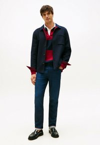 Joven de pie vistiendo una chaqueta azul marino, una camiseta de rugby a rayas rojas y azul marino, jeans azul oscuro y mocasines de cuero negros sobre un fondo blanco.