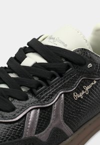 Μαύρο αθλητικό παπούτσι με επάνω μέρος από δέρμα με υφή, λευκή γλώσσα, μαύρα κορδόνια και ασημί λεπτομέρειες· διαθέτει το χαρακτηριστικό λογότυπο στη πλευρά.