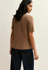 Brązowy sweter z krótkim rękawem z ribbowanym wykończeniem u dołu i na mankietach, wykonany z miękkiego materiału. W połączeniu z ciemnoniebieskimi spodniami o szerokich nogawkach. Widok z tyłu.