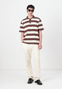 Hnedo-biely pruhovaný polo tričko s golierom, skombinované s smotanovými nohavicami a čiernymi topánkami. Model má na sebe tmavé slnečné okuliare.