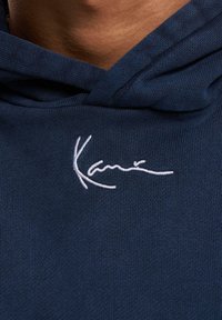 Sudadera azul marino de textura suave. Cuenta con un logo de firma bordado en blanco en el área del pecho. Capucha con cordón ajustable.