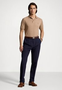 Μπεζ πόλο μπλούζα, ναυτικό μπλε παντελόνι με λεπτές ρίγες, καφέ δερμάτινη ζώνη και μοκασίνια. Κλασική εφαρμογή με κοντά μανίκια και διακριτικό λογότυπο στην μπλούζα.