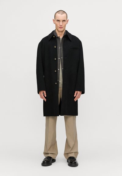 Melnas vilnas mētelis ar sakrautām apkaklēm, pogu slēgšanu un krūšu kabatu. Slim fit ar garām piedurknēm, saskaņots ar bēšiem biksēm un melniem apaviem.