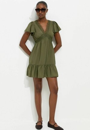 Donna che indossa occhiali da sole scuri, un vestito corto verde oliva con volant e maniche svolazzanti, e ballerine nere intrecciate, in piedi su sfondo bianco.