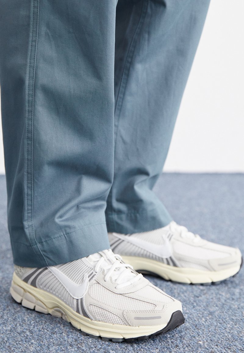 Białe sportowe sneakersy z teksturowanej siateczki i gumowymi podeszwami, z szarymi akcentami. Noszone z jasnoniebieskimi, lekko luźnymi spodniami.