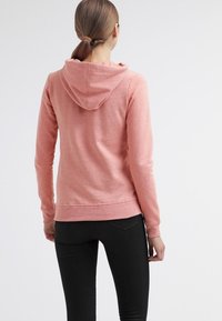 Mujer con el cabello castaño recogido, vistiendo una sudadera con capucha rosa y pantalones negros, de espaldas contra un fondo claro y liso.