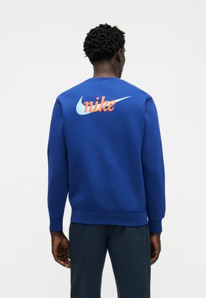 Hombre vistiendo una sudadera azul con el logo de Nike en azul claro y texto naranja "nike" en la espalda, de pie frente a un fondo claro y liso.